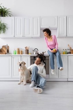 Smiling african american woman holding orange juice near boyfriend with labrador at home 