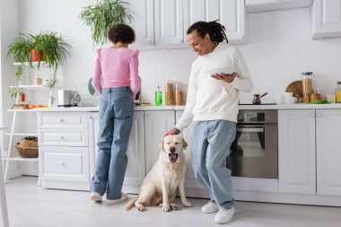 African american woman washing dishes near boyfriend petting labrador at home 