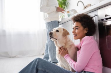 Smiling african american woman hugging labrador near blurred boyfriend at home 