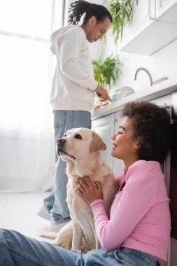 Side view of smiling african american woman hugging labrador near blurred boyfriend in kitchen 