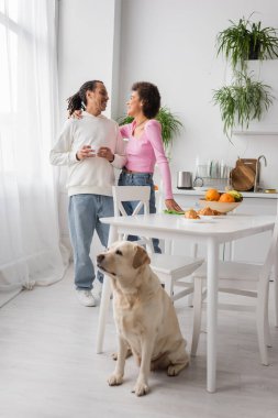 African american woman hugging boyfriend with coffee near labrador in kitchen 