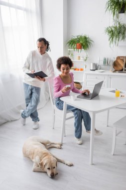 African american freelancer using devices near boyfriend with notebook and labrador at home 