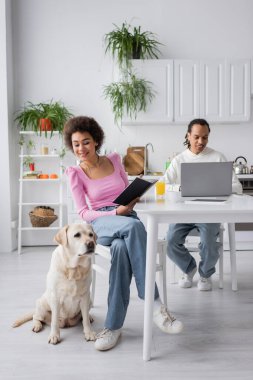 African american woman holding notebook near labrador and boyfriend using laptop at home 