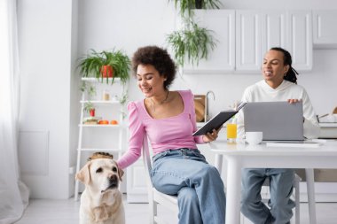 African american woman holding notebook and petting labrador near boyfriend and devices at home 