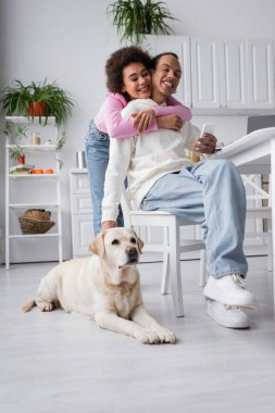 African american woman hugging boyfriend with smartphone near labrador at home 