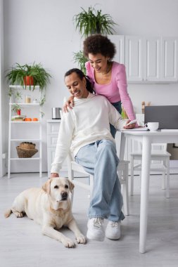 Smiling african american woman hugging boyfriend near labrador and devices in kitchen 