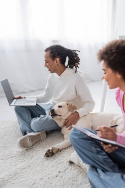 Side view of labrador lying near african american freelancers working on carpet at home  clipart