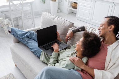 Blurred african american couple using laptop with blank screen while lying on couch at home 