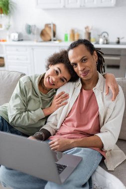 Smiling african american woman looking at camera near laptop on couch at home 