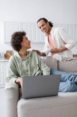 Positive african american couple with coffee and laptop talking at home 