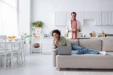 Positive african american couple with laptop and coffee looking at camera in kitchen 