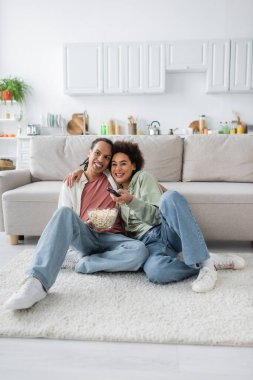 Smiling african american woman hugging boyfriend with popcorn while watching tv at home  clipart