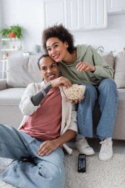 African american woman holding popcorn near boyfriend and remote controller at home 