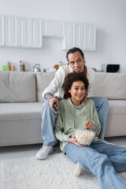 Gülümseyen Afrikalı Amerikalı çift film izliyor ve evde patlamış mısır tutuyorlar. 