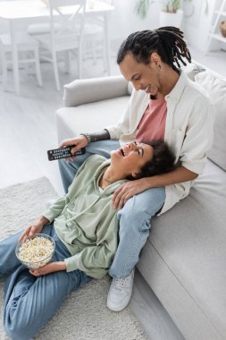 Positive african american couple holding remote controller and popcorn at home 