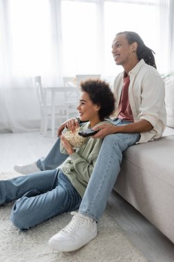 Cheerful african american couple holding remote controller and popcorn at home 