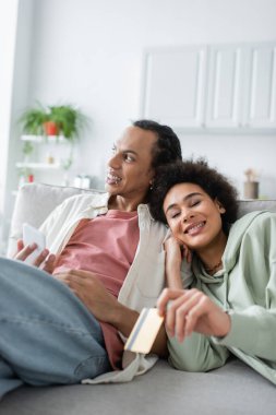 Smiling african american woman holding blurred credit card near boyfriend with mobile phone at home 