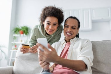 Gülümseyen Afrikalı Amerikalı çift evde online alışveriş yaparken akıllı telefon kullanıyor. 