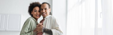 African american couple hugging and looking at camera in kitchen, banner 