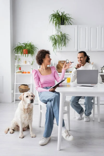 Smiling african american woman with notebook talking to boyfriend near devices and labrador at home