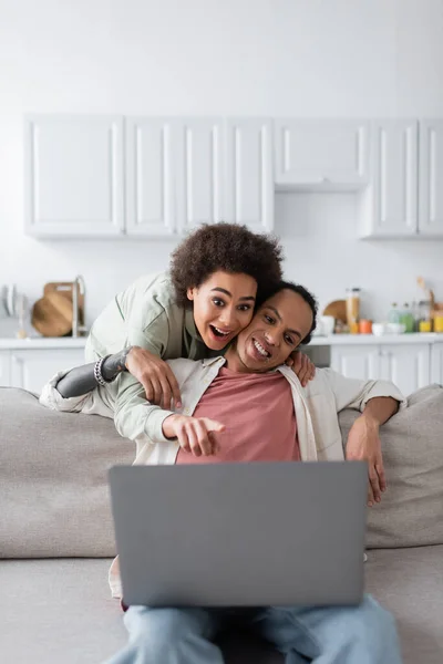 Excited african american woman looking at blurred laptop near boyfriend on couch at home