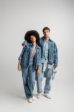 full length of young man in denim outfit hugging african american woman on grey 