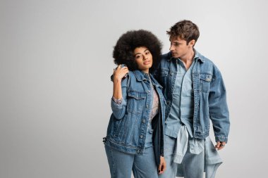 young man in denim outfit hugging curly african american woman isolated on grey 