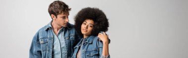 young man in denim outfit hugging curly african american woman isolated on grey, banner 