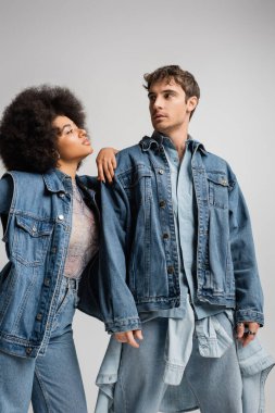 curly african american model leaning on shoulder of young man in denim outfit isolated on grey 