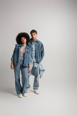 full length of young interracial man and woman standing in total denim outfit on grey 