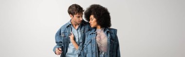 curly african american model touching denim jacket of young man isolated on grey, banner 