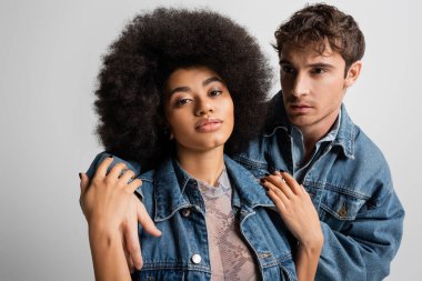 man in denim outfit hugging curly african american woman isolated on grey 