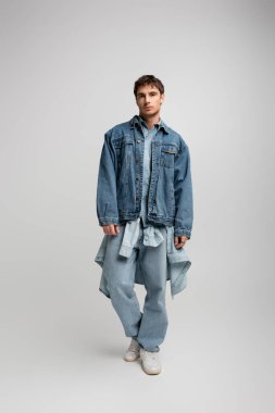 full length of young man in denim outfit posing and looking at camera on grey 