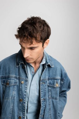 young man in denim jacket looking away while posing isolated on grey 