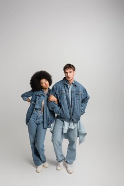 full length of young man in denim outfit posing with hands in pockets near curly african american woman on grey  clipart