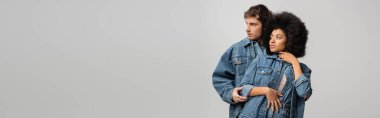 young multiethnic couple in denim outfit hugging and looking away isolated on grey, banner 
