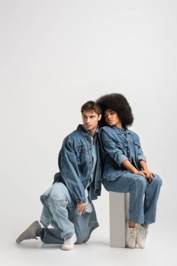 full length of african american woman sitting near man in denim outfit on grey 