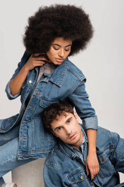 brunette african american woman sitting and looking at man in denim outfit isolated on grey 