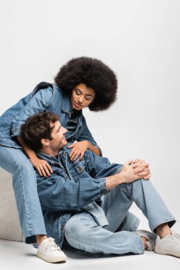brunette african american woman in sneakers sitting and looking at cheerful man in denim outfit on grey 