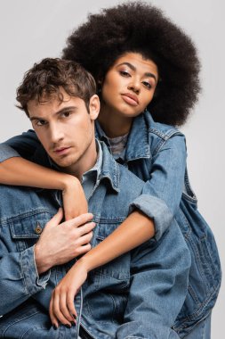 brunette african american model looking at camera and hugging stylish man isolated on grey 