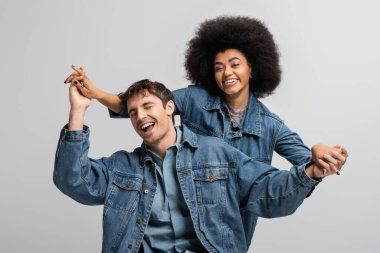 cheerful african american model looking at camera and holding hands with stylish man isolated on grey 