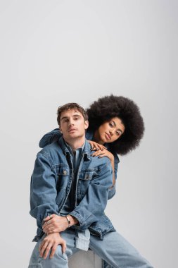 curly african american model looking at camera with stylish man sitting isolated on grey 