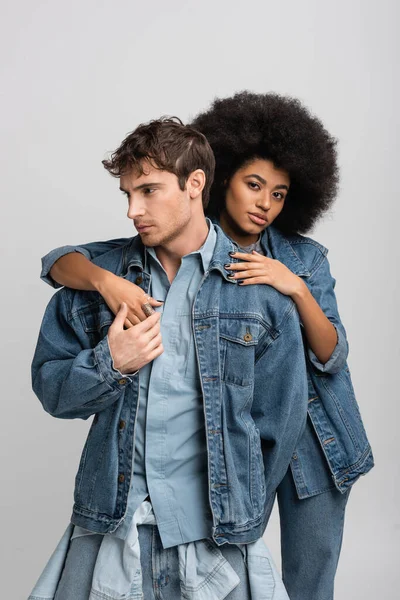 curly african american model looking at camera while embracing stylish man in denim clothes isolated on grey