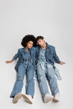 full length of stylish interracial couple in denim clothes sitting in sneakers on grey 