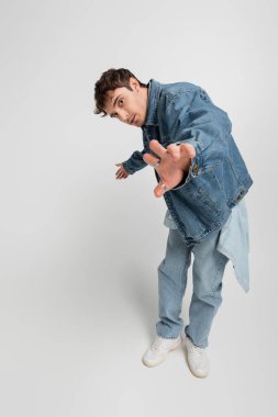 full length of stylish young man in denim jacket and white sneakers standing with outstretched hand on grey 