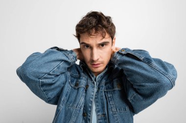 stylish young man in denim jacket posing and looking at camera isolated on grey 