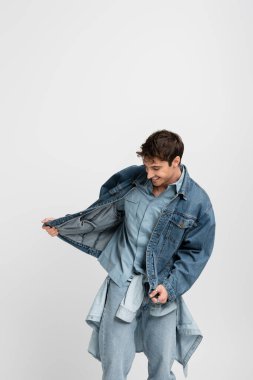 happy young man in denim outfit posing while adjusting jacket isolated on grey 