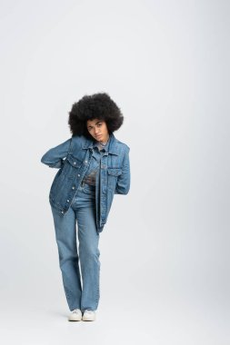 full length of curly african american woman in denim outfit looking at camera while standing on grey 