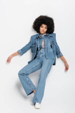 full length of curly african american woman in denim jacket looking at camera while posing near grey wall 