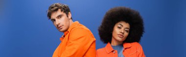 Multiethnic couple in orange jackets looking at camera isolated on blue, banner 